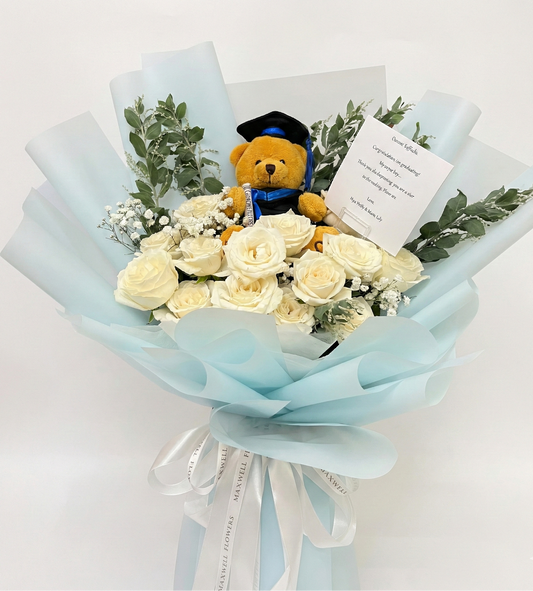 Bouquet of white flowers with a teddy bear in a graduation cap on a light blue wrap against a white background.