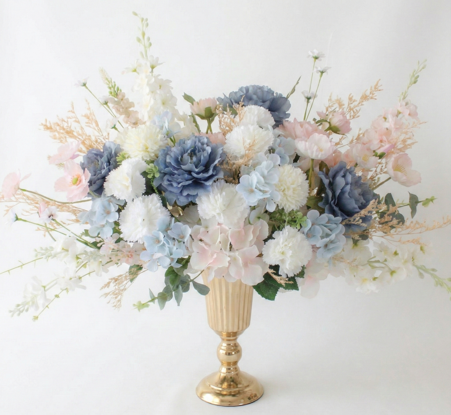 Floral arrangement with blue, white, and pink flowers in a gold vase on a white background