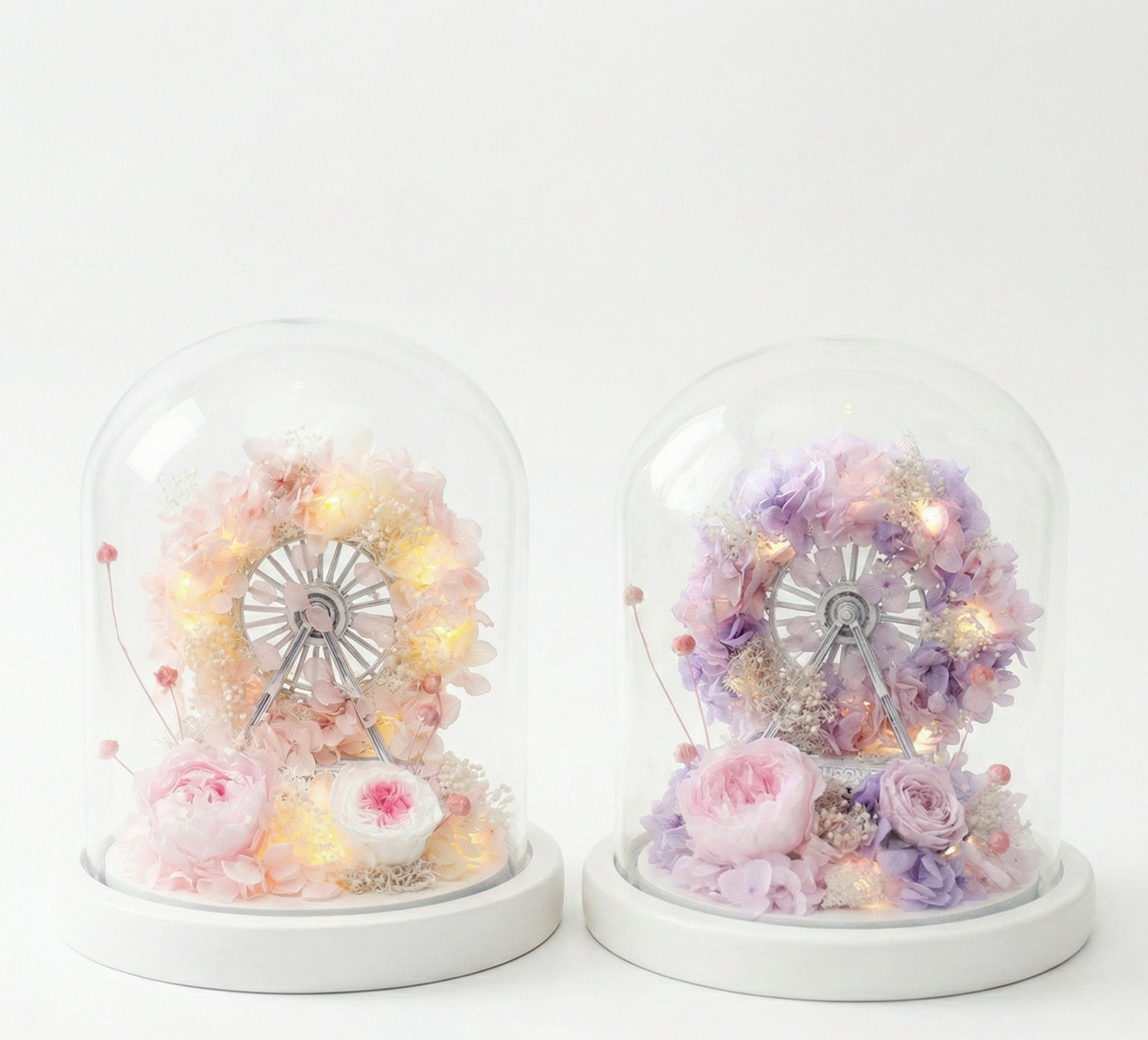 Two decorative items with floral arrangements and a Ferris wheel inside glass domes on a white background.